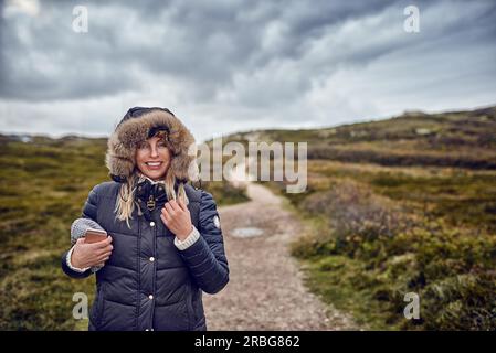 Frau mittleren Alters braving kalten Winter Tag in die Natur zu Fuß durch die Landschaft an einem breezy Tag glücklich in die Kamera lächeln Stockfoto