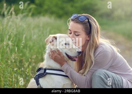 Fröhlich lächelnder goldener Hund, der ein Gurt trägt, sitzt mit Blick auf seine hübsche junge Besitzerin, die ihn draußen mit einem liebevollen Lächeln streichelt Stockfoto