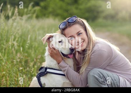 Fröhlich lächelnder goldener Hund, der ein Gurt trägt, sitzt mit Blick auf seine hübsche junge Besitzerin, die ihn draußen mit einem liebevollen Lächeln streichelt Stockfoto