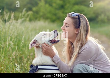 Fröhlich lächelnder goldener Hund, der ein Gurt trägt, sitzt mit Blick auf seine hübsche junge Besitzerin, die ihn draußen mit einem liebevollen Lächeln streichelt Stockfoto