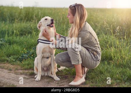 Fröhlich lächelnder goldener Hund, der ein Gurt trägt, sitzt mit Blick auf seine hübsche junge Besitzerin, die ihn draußen mit einem liebevollen Lächeln streichelt Stockfoto