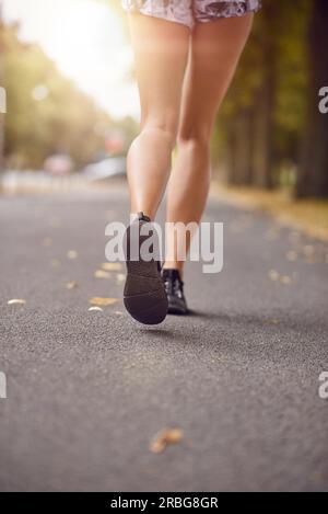 Eine junge Frau geht oder joggt eine Herbststraße hinunter, die vom Schein der Abendsonne in einem niedrigen Winkel auf ihre formschönen Beine in kurzen Hosen und beleuchtet wird Stockfoto