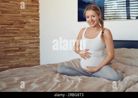 Glücklich schwanger Frau sitzt auf einem Bett mit Ihrem Babybauch mit ihren Händen in die Kamera lächeln Stockfoto