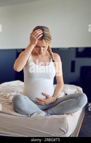 Schwer schwangere junge Frau, die quer auf einem Bett sitzt, ihren geschwollenen Unterleib umklammert und ihren Kopf mit einem ernsten Blick nach unten umklammert Stockfoto