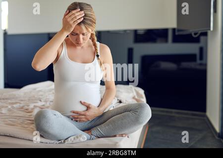 Schwer schwangere junge Frau, die quer auf einem Bett sitzt, ihren geschwollenen Unterleib umklammert und ihren Kopf mit einem ernsten Blick nach unten umklammert Stockfoto