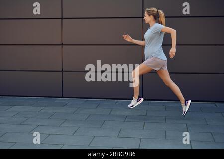 Schwangere junge Frau fit halten Sprinten entlang eines gepflasterten Gehweg vor einer Wand in einem gesunden Lebensstil Konzept Stockfoto