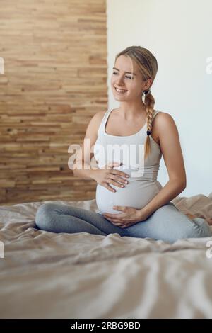 Glückliche, schwangere Frau, die sich mit ihrem ungeborenen Kind verbindet, sitzt auf einem Bett und wiegt ihr Baby mit ihren Händen, die seitlich mit einem zärtlichen Lächeln schauen Stockfoto