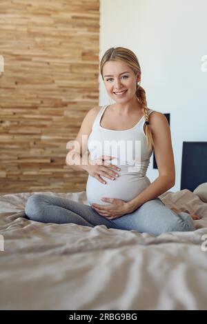 Glücklich schwanger Frau sitzt auf einem Bett mit Ihrem Babybauch mit ihren Händen in die Kamera lächeln Stockfoto
