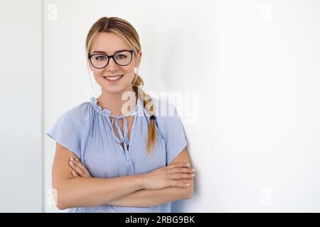 Freundliche junge blonde Frau, die eine Brille trägt, deren langes Haar über einer Schulter geflochten ist, und die Arme gefaltet, die die Kamera anlächelt und sich auf ein Weiß lehnt Stockfoto