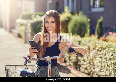 Wunderschöne junge Frau mit langen Haaren in einem halblangen Porträt draußen in der Stadt, die sich mit einem modernen Smartphone auf ein altes Fahrrad lehnt Stockfoto