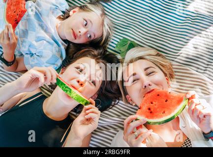 Mutter liegt mit Töchtern auf einer Picknickdecke im Stadtpark an sonnigen Wochenendtagen, lächelt, lacht vor der Kamera und isst saftige Wassermelone. Fa Stockfoto