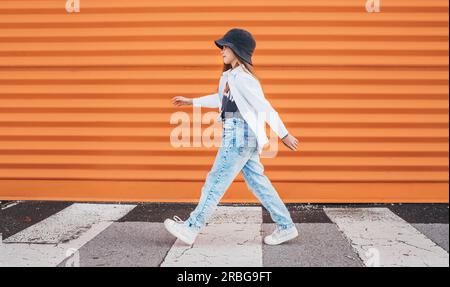 Kleine Mode-gekleidete Mädchen lustig zu Fuß über einen Fußgängerzebra Crosswalk auf der orangefarbenen Wand Hintergrund. Urbane Menschen leben und eine Straße jeden Stockfoto