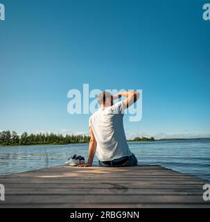 Ein Mann, der im Sommer in Finnland auf einem hölzernen Steg am See sitzt Stockfoto