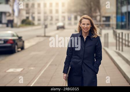 Attraktive stilvolle blonde Frau verlassen Ihren Arbeitsplatz als sie Spaziergänge eine ruhige high key Urban Street lächelnd, als sie ihren Laptop trägt mit Kopie Raum Stockfoto