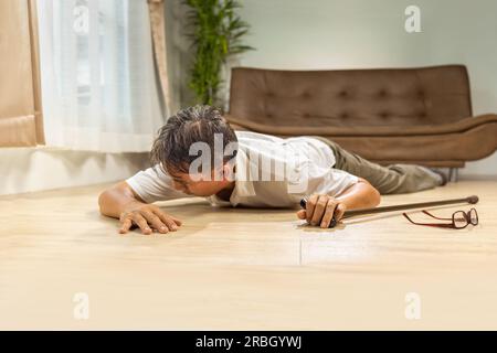 Asiatischer Senior-Mann fiel hinunter und lag auf dem Boden, während er zu Hause mit Gehstock spazieren ging. Stockfoto