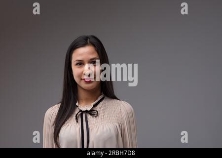 Ein halblanges Porträt einer stolzen, intelligenten, selbstbewussten, jungen Filipina-Frau. Sie trägt eine beige Bluse mit schwarzem Band. Grauer Hintergrund Stockfoto