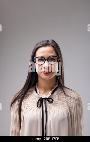 Porträt einer selbstbewussten, intelligenten Filipina-Frau mit langen Haaren. Mit Brille. Gekleidet in einer beigen Bluse mit schwarzem Band. Grauer Hintergrund Stockfoto