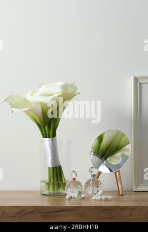 Wunderschöne Calla Lilien Blumen in Glasvase, Flaschen mit Parfüm, Schmuck und Spiegel auf Holztisch Stockfoto