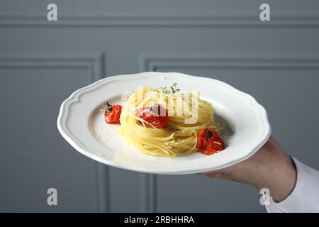 Der Kellner hielt einen Teller mit leckerem Capellini mit Tomaten und Käse in der Nähe der grauen Wand, Schließung. Exquisite Präsentation eines Nudelgerichts Stockfoto