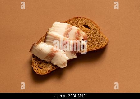 Eine Scheibe Schwarzbrot und Speck liegt auf braunem Hintergrund, Brot mit Speck Stockfoto