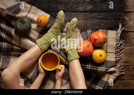 Ein Mädchen sitzt in Socken auf dem Boden und hält eine Tasse Tee auf einer Decke und Kürbisse liegen in der Nähe im Herbst, zu Hause und bequem, trinken Tee im Herbst, fe Stockfoto