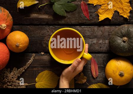 Die weibliche Hand hält eine Tasse Tee neben Kürbis und Herbstblättern auf dem Tisch, Herbsttisch mit einer Tasse Tee in den Händen, Herbststimmung Stockfoto