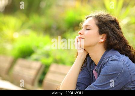 Eine Frau, die sich entspannt und die Augen schließt, sitzt auf einer Bank im Park Stockfoto