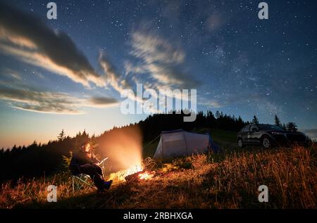 Camping in den Bergen unter bewölktem Nachthimmel. Junger Mann, der sich am Lagerfeuer ausruht, in der Nähe eines Touristenzelts und eines Geländewagens auf einem Hügel. Ein reisender Mann, der nachts die Sterne beobachtet. Stockfoto