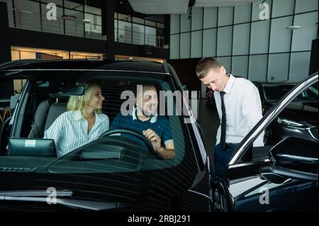Der Verkäufer demonstriert den Käufern das Fahrzeuginnere. Ein weißes Paar sucht sich ein Auto aus. Stockfoto