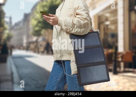 Unerkennbare Frau mit flexibler Solarbatterie, die das Smartphone im Hintergrund einer Stadt hält. Reise- und Alternativenergiekonzept. Stockfoto