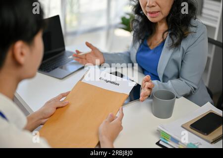Nahaufnahme eines jungen, männlichen Büromitarbeiters in Asien, der ein ernstes Gespräch führt und ein Kündigungsschreiben an eine leitende Chefin im Büro übergibt. Stockfoto