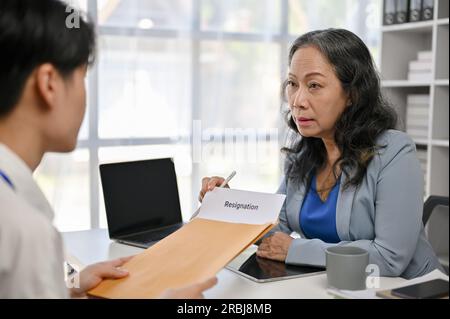 Ein junger, männlicher Büroangestellter aus Asien hält ein ernstes Gespräch und gibt einem weiblichen Chefin im Büro ein Kündigungsschreiben. Stockfoto