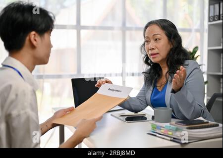 Ein junger, männlicher Büroangestellter aus Asien hält ein ernstes Gespräch und gibt einem weiblichen Chefin im Büro ein Kündigungsschreiben. Stockfoto