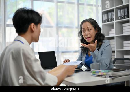 Ein junger, männlicher Büroangestellter aus Asien hält ein ernstes Gespräch und gibt einem weiblichen Chefin im Büro ein Kündigungsschreiben. Zurücktreten, einen Job kündigen, Stockfoto