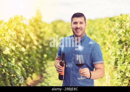 Lächelnder junger Mann, der ein Glas und eine Flasche Wein auf dem Weinberg hielt Stockfoto