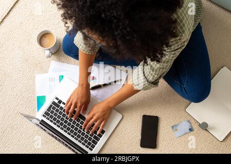 Blick von oben auf eine birassische Frau mit Laptop, Smartphone und Kreditkarte, die zu Hause auf dem Boden sitzt. Lebensstil, Kommunikation und häusliches Leben, unveränderlich Stockfoto