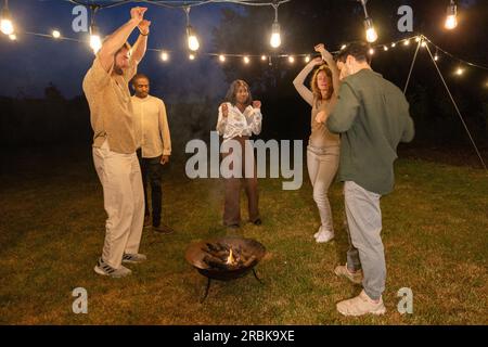 Mehrere ethnische Gruppen von Freunden, die im Garten grillen, lachen und tanzen und einer Gruppe von Freunden zeigen, wie sie sich mit den Sparklern am Abend amüsieren. Hochwertiges Foto Stockfoto