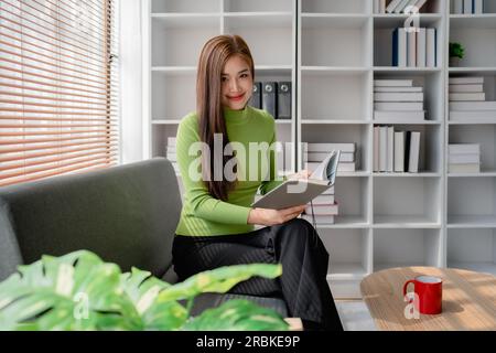 Eine junge asiatische Frau, die sich Notizen in Papierheft macht, sitzt auf einem gemütlichen Sofa im Hintergrund des Fensters. Hübsche Frau schreibt Notizen in Tagebüchern oder macht To-Do Stockfoto