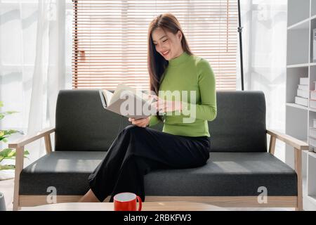 Eine junge asiatische Frau, die sich Notizen in Papierheft macht, sitzt auf einem gemütlichen Sofa im Hintergrund des Fensters. Hübsche Frau schreibt Notizen in Tagebüchern oder macht To-Do Stockfoto