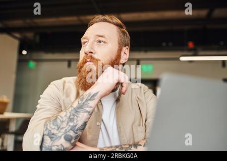 Porträt eines nachdenklichen, tätowierten und bärtigen Unternehmers, der am Arbeitsplatz sitzt, wegschaut und in der Nähe des Laptops denkt, auf verschwommenem Vordergrund in der modernen Version Stockfoto
