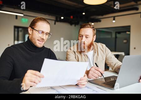 Lächelnder Unternehmer in schwarzem Rollkragen und einer Brille, der in der Nähe eines überraschten, bärtigen Geschäftspartners auf Papier schaute, Karten und Laptop auf dem Schreibtisch in CO Stockfoto