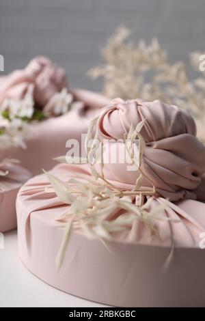 Furoshiki-Technik. Geschenke in pinkfarbenem Stoff und getrockneten Zweigen auf weißem Tisch, Nahaufnahme Stockfoto