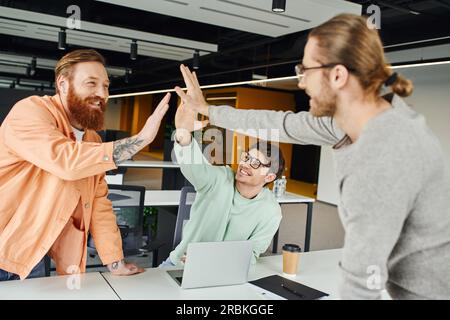 Aufgeregtes Business-Team, mit Bart tätowierter Mann und Kollegen mit Brille geben High Five und bestätigen die Zustimmung in der Nähe des Laptops in moderner Zusammenarbeit Stockfoto