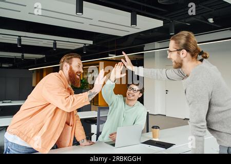 Ein überfreulicher, bärtiger und tätowierter Unternehmer gibt High Five an Geschäftspartner in der Nähe eines Laptops und ein Getränk zum Mitnehmen in einer modernen Umgebung der Zusammenarbeit von Stockfoto