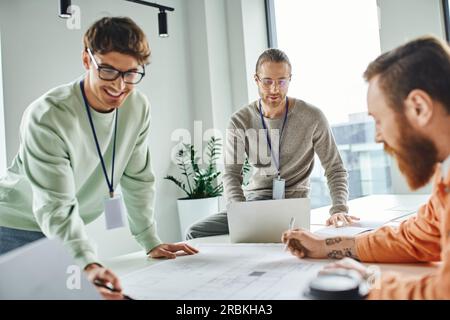 Ein lächelnder Architekt, der auf einen Plan in der Nähe von Kollegen und Laptops zeigt, während er in einem Architekturstudio, einer Geschäftszusammenarbeit und Stockfoto