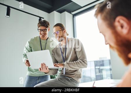 Positive Emotionen, erfolgreiche und lächelnde Designer in einer Brille, die auf ein Notebook in der Nähe eines Kollegen schauen, der in einer modernen Zusammenarbeit an verschwommenem Vordergrund arbeitet Stockfoto