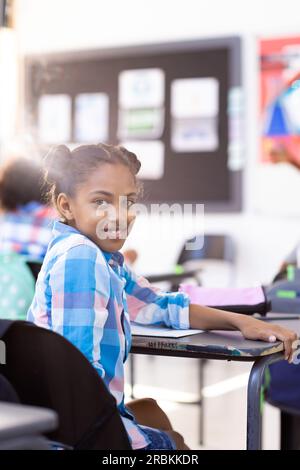 Vertikales Porträt eines lächelnden, birassischen Schulmädchens, das am Schreibtisch sitzt und sich im Klassenraum umdreht. Bildung, Inklusivität, Grundschule, Lernen Stockfoto