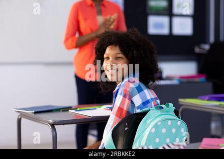 Porträt eines lächelnden, birassischen Schulmädchens, das am Schreibtisch sitzt und sich im Klassenraum umdreht. Bildung, Inklusivität, Grundschule, Lernkonzept Stockfoto