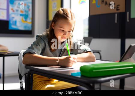 Ein weißes Schulmädchen sitzt am Schreibtisch und macht sich Notizen im Klassenzimmer Stockfoto