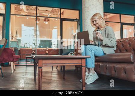 Geschäftsfrau mittleren Alters, die mit einem Laptop arbeitet und Kaffee trinkt, in gemütlicher Zusammenarbeit Stockfoto